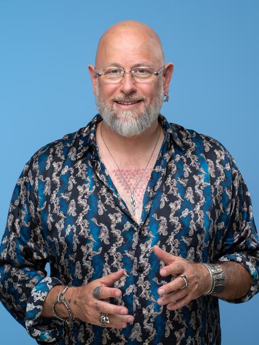 Porträt des Hypnotiseurs und Bühnenkünstlers Jan Becker. Er trägt ein blau-gemustertes Hemd und viel Schmuck und Tätowierungen. Der Hintergrund ist hell-blau. Er guckt in die Kamera. 
