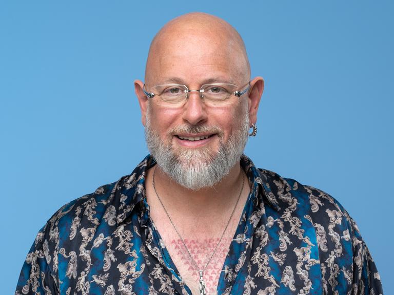 Porträt des Hypnotiseurs und Bühnenkünstlers Jan Becker. Er trägt ein blau-gemustertes Hemd und viel Schmuck und Tätowierungen. Der Hintergrund ist hell-blau. Er guckt in die Kamera. 