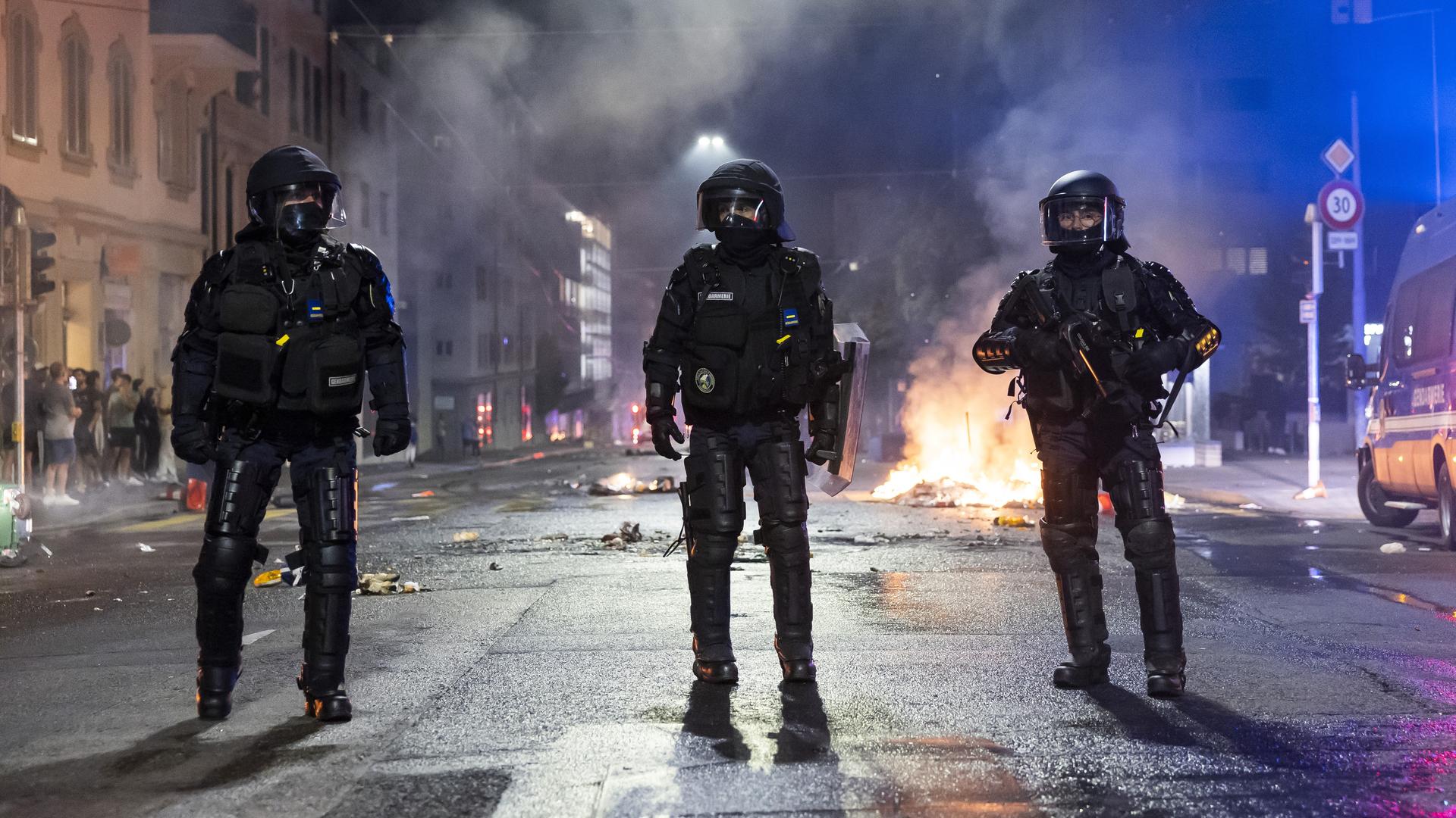 Es ist dunkel. Drei Polizisten in voller Kampfmonitur stehen nebeneinander auf einer Straße. Dahinter sieht man ein Feuer und Trümmer auf der Straße sowie eine Häuserwand. Rauch steigt auf. Es ist dunkel. Drei Polizisten in voller Kampfmonitur stehen nebeneinander auf einer Straße. Dahinter sieht man ein Feuer und Trümmer auf der Straße sowie eine Häuserwand. Rauch steigt auf.