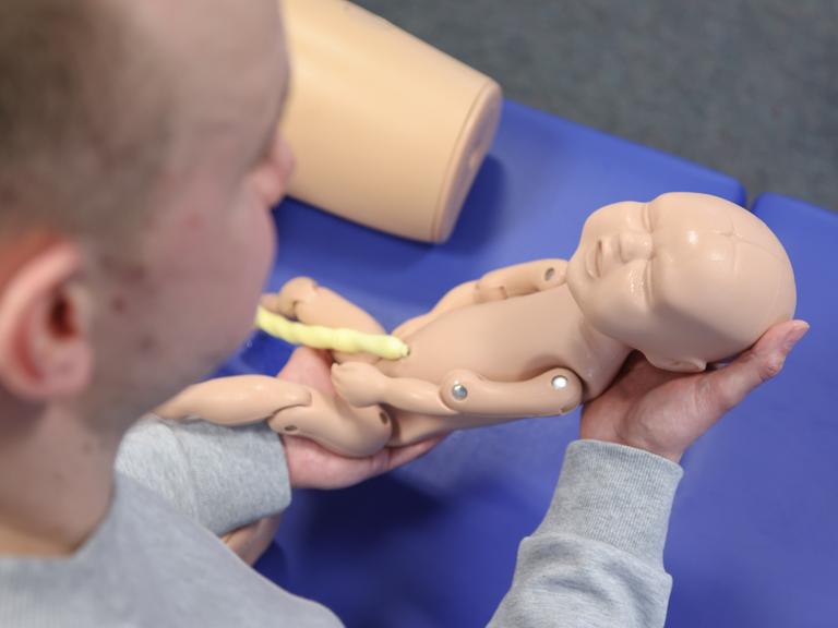 Eine männliche Hebamme, hält in einer gestellten Situation eine Neugeborenen-Übungspuppe mit Nabelschnur. Eine männliche Hebamme, hält in einer gestellten Situation eine Neugeborenen-Übungspuppe mit Nabelschnur.