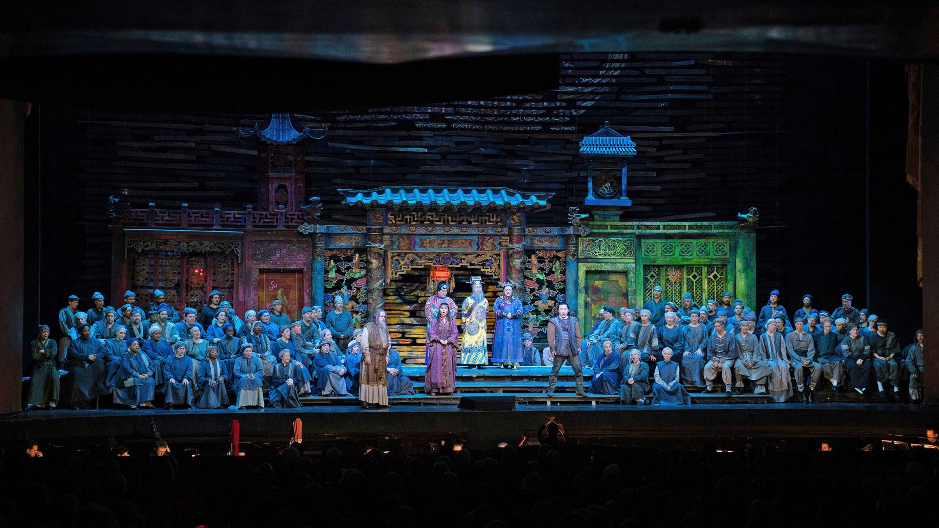 Auf der Bühne der Metropolitan Opera in New York einer "Turandot"-Inszenierung stehen dutzende Sänger und Darsteller in blauen Kostümen vor der Kulisse eines Hauses mit Treppe.