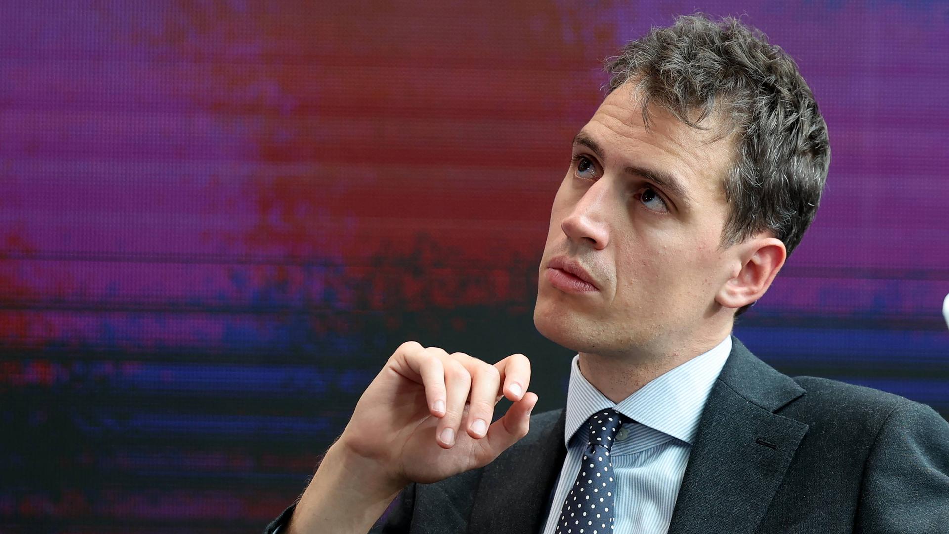 Arthur Mensch in Anzug und Krawatte, schaut nach rechts oben. Im Hintergrund sind lediglich Farben eines großen Bildschirms zu erkennen.