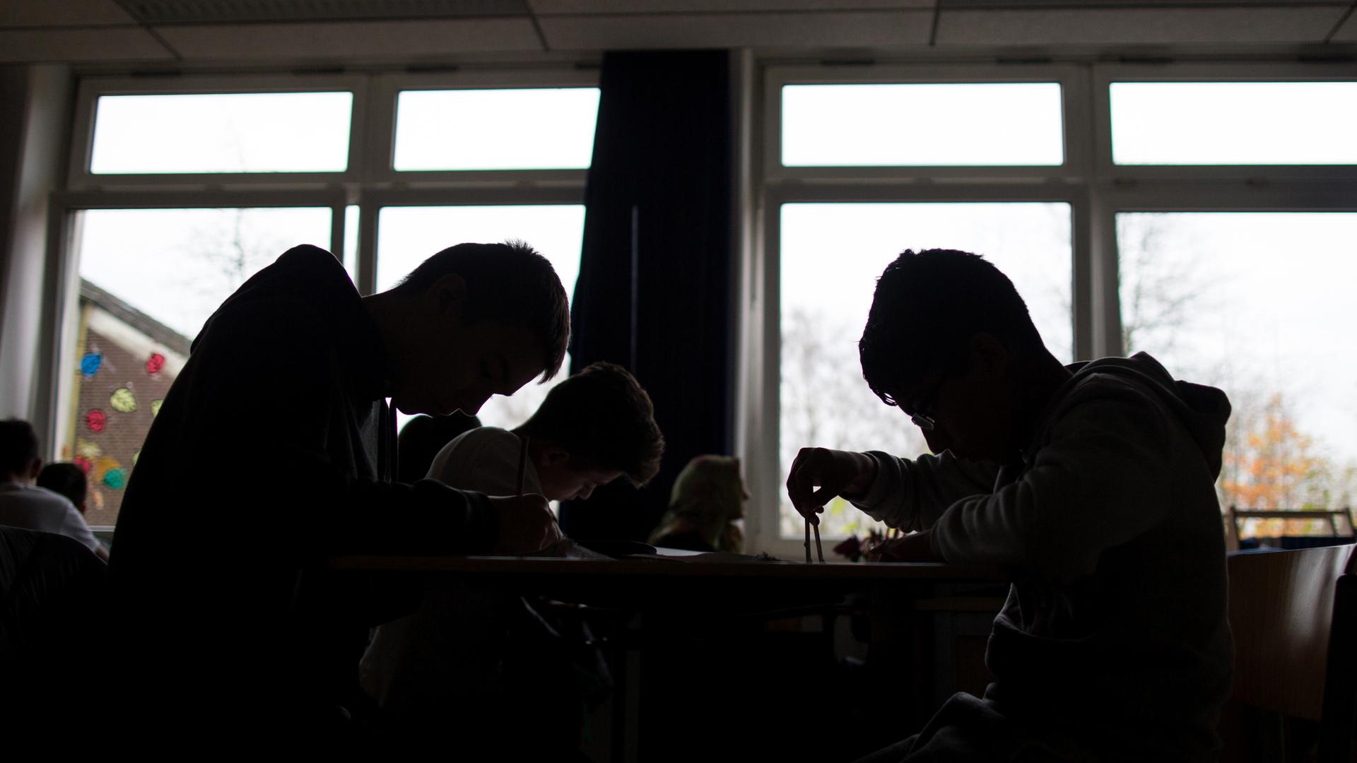 Schüler sitzen während einer Unterrichtsstunde arbeitend an Tischen. Sie heben sich als dunkle Silhouetten vor den Fenstern ab. Schüler sitzen während einer Unterrichtsstunde arbeitend an Tischen. Sie heben sich als dunkle Silhouetten vor den Fenstern ab.