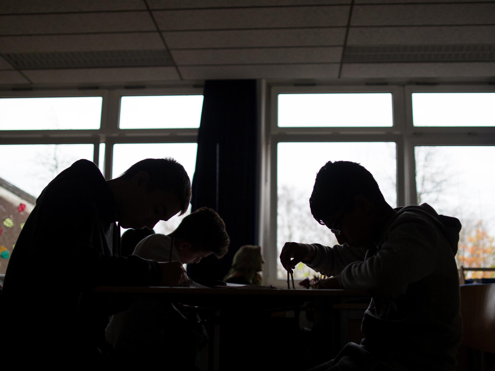 Schüler sitzen während einer Unterrichtsstunde arbeitend an Tischen. Sie heben sich als dunkle Silhouetten vor den Fenstern ab.