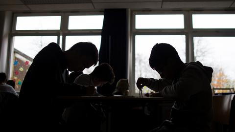 Schüler sitzen während einer Unterrichtsstunde arbeitend an Tischen. Sie heben sich als dunkle Silhouetten vor den Fenstern ab.