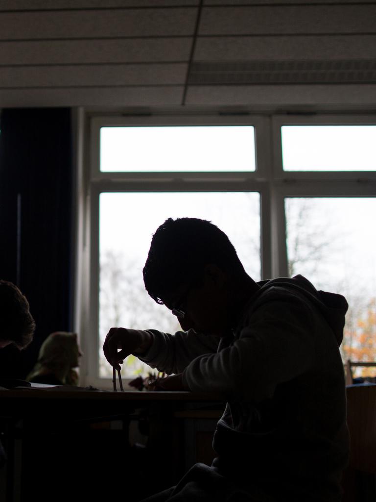 Schüler sitzen während einer Unterrichtsstunde arbeitend an Tischen. Sie heben sich als dunkle Silhouetten vor den Fenstern ab.