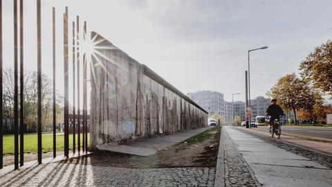 Auf einer Straße sind Reste der Berliner Mauer zu sehen. An einer Stelle stehen nur noch die stützenden Stahlstangen, sodass die Mauer durchlässig ist.