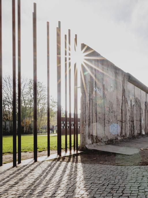 Auf einer Straße sind Reste der Berliner Mauer zu sehen. An einer Stelle stehen nur noch die stützenden Stahlstangen, sodass die Mauer durchlässig ist.