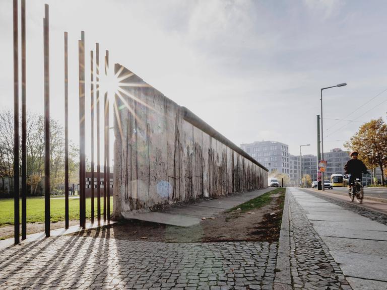 Auf einer Straße sind Reste der Berliner Mauer zu sehen. An einer Stelle stehen nur noch die stützenden Stahlstangen, sodass die Mauer durchlässig ist.