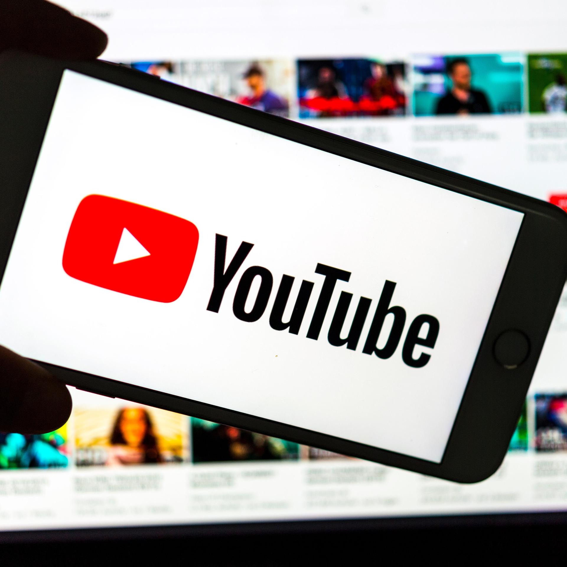 Das Logo des Video-Portals YouTube wird auf dem Display eines Smartphones angezeigt. Im Hintergrund ist auf einem Bildschirm die YouTube Homepage zu sehen.