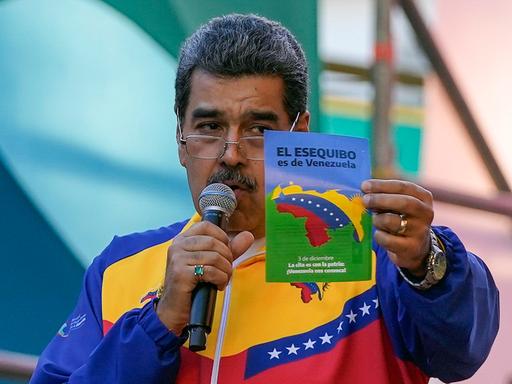 Maduro trägt eine Jacke in Farben der venezolanischen Flagge, spricht in ein Mikrophon, das er in der Hand hält, und zeigt mit der anderen Hand einen Flyer hoch, der Venezuelas Anspruch auf die Region Essequibo erhebt.