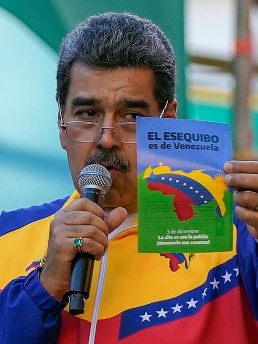 Maduro trägt eine Jacke in Farben der venezolanischen Flagge, spricht in ein Mikrophon, das er in der Hand hält, und zeigt mit der anderen Hand einen Flyer hoch, der Venezuelas Anspruch auf die Region Essequibo erhebt.