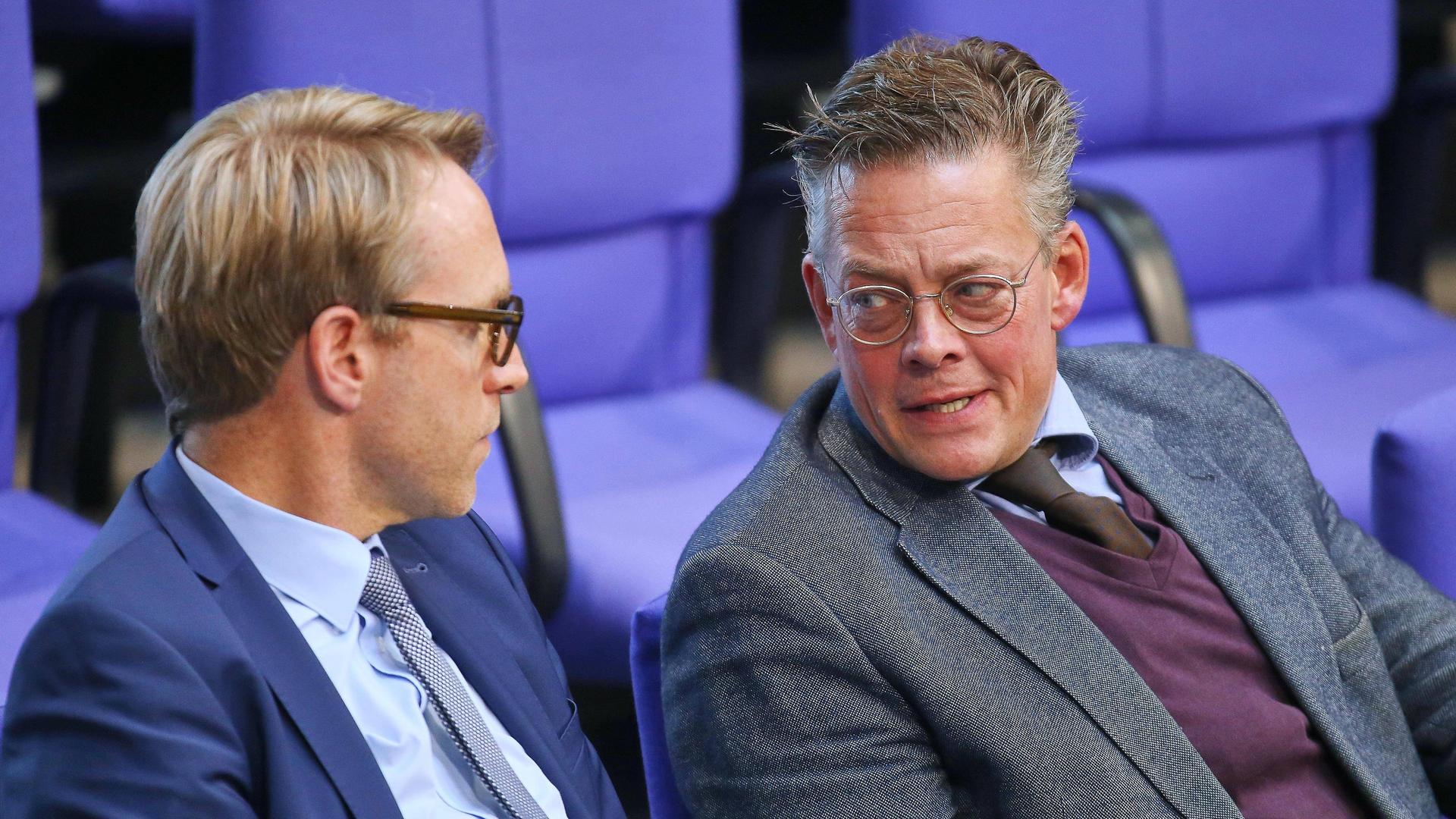 Marc Henrichmann und Konstantin von Notz sitzen nebeneinander im Plenum des Bundestags und reden miteinander.