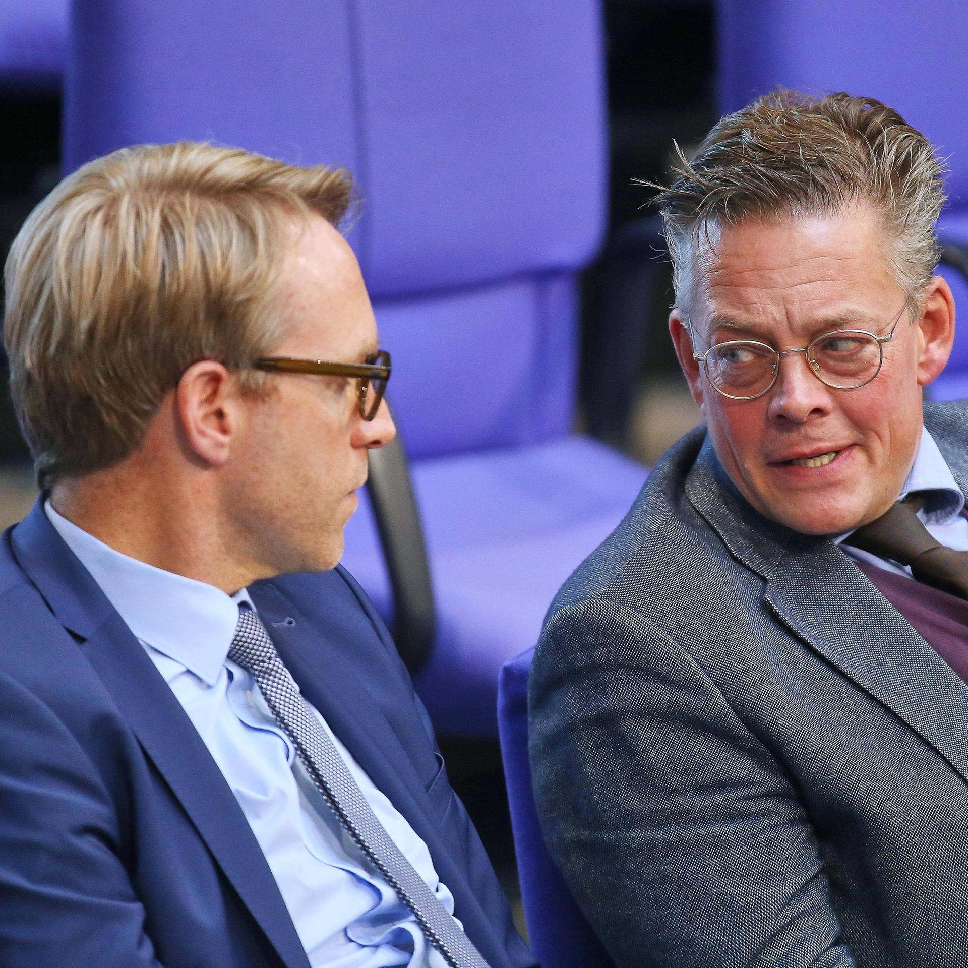 Marc Henrichmann und Konstantin von Notz sitzen nebeneinander im Plenum des Bundestags und reden miteinander.