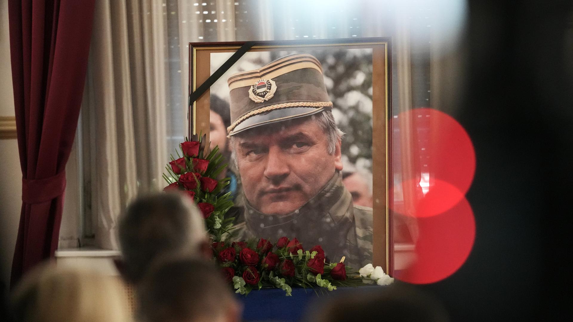 Ein Porträt des bosnisch-serbischen Kriegsverbrechers Ratko Mladić in Uniform ist in einem Rahmen aufgestellt. Vor dem Rahmen ist ein Bouquet roter Rosen angebracht. 