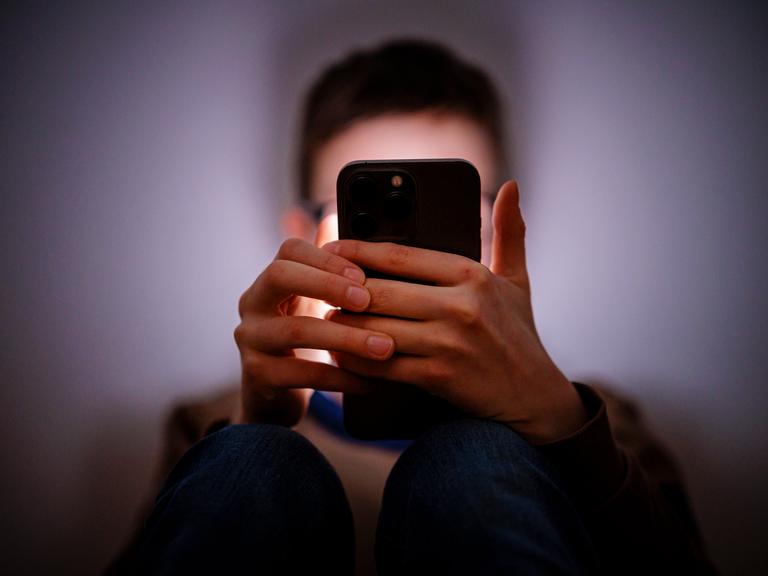 Ein Junge sitzt in einem dunklen Raum und wird von einem Smartphone beleuchte, das er mit beiden Händen vor sein Gesicht hält. (Symbolfoto)