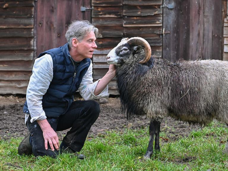 Holger Siemann, Bauer, Schriftsteller und Hörspielautor, hockt in seinem Garten in der Uckermark vor einem Gotlandschaf. 