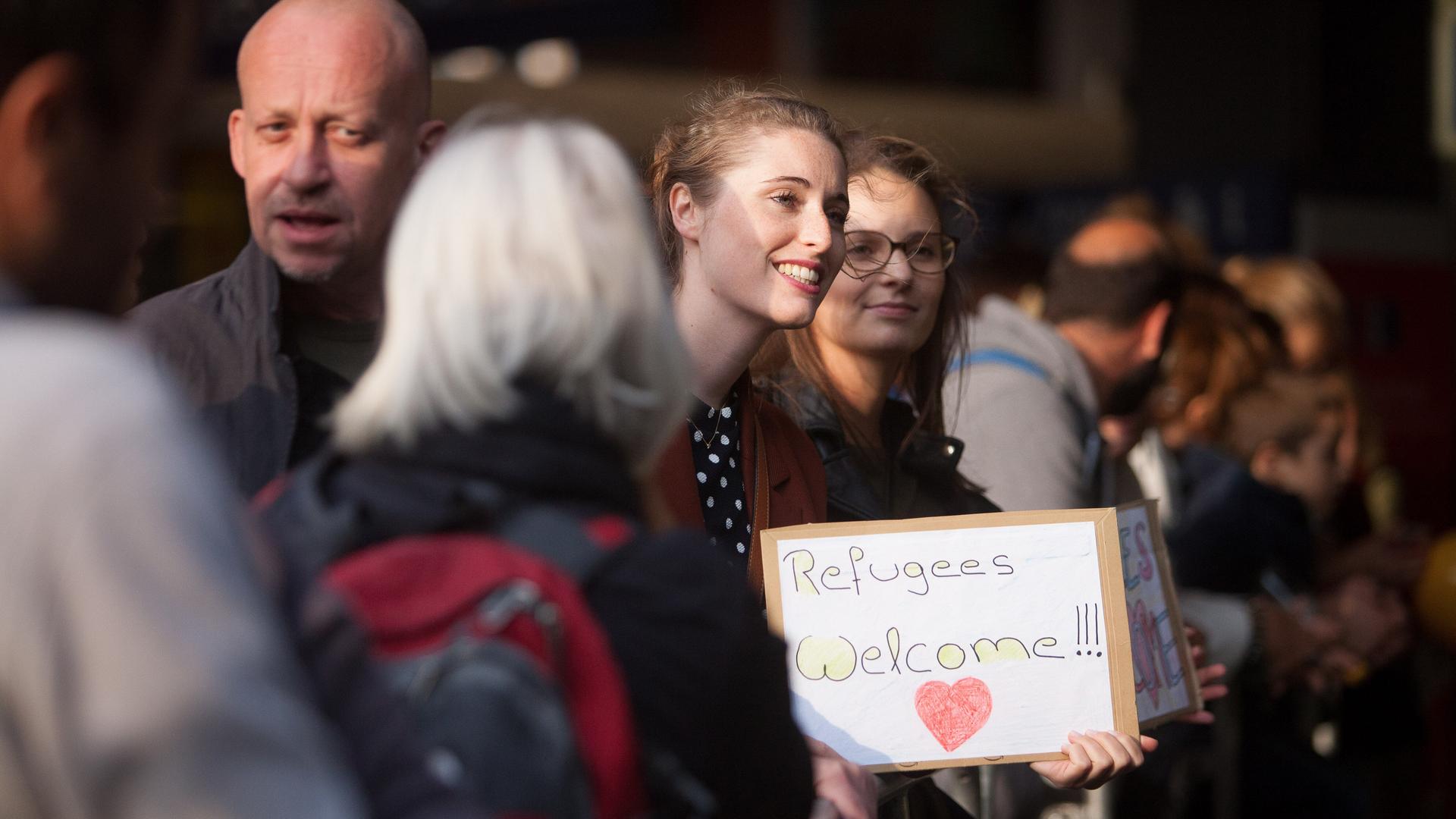 Menschen stehen 2015 Hauptbahnhof in München, eine Frau hält ein Schild mit der Aufschrift "Refugees Welcome" in den Händen. Menschen stehen 2015 Hauptbahnhof in München, eine Frau hält ein Schild mit der Aufschrift "Refugees Welcome" in den Händen.