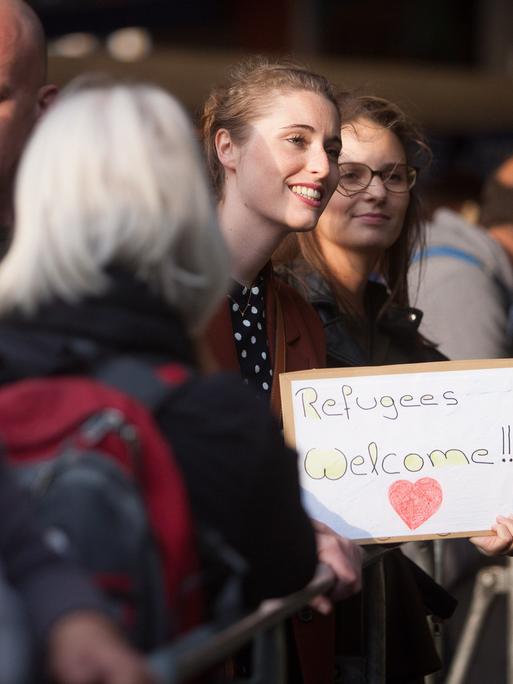 Menschen stehen 2015 Hauptbahnhof in München, eine Frau hält ein Schild mit der Aufschrift "Refugees Welcome" in den Händen.
