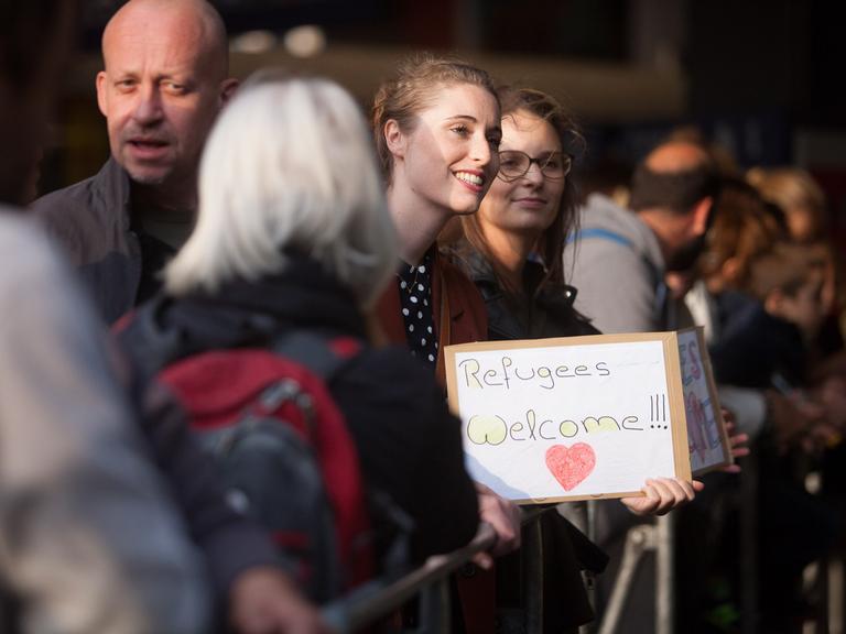 Menschen stehen 2015 Hauptbahnhof in München, eine Frau hält ein Schild mit der Aufschrift "Refugees Welcome" in den Händen.