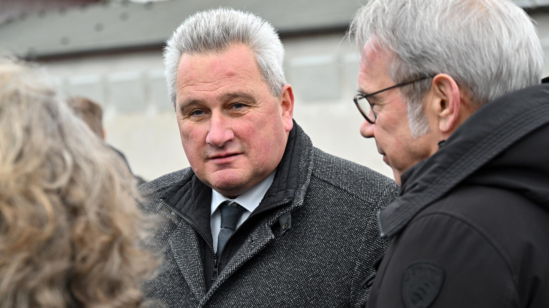 Jens-Christian Wagner, Direktor der Stiftung Gedenkstätten Buchenwald und Mittelbau-Dora, steht auf dem Appellplatz in der Gedenkstätte Buchenwald zwischen zwei Personen und schaut in die Kamera. Er trägt einen dicken grauen Wintermantel.