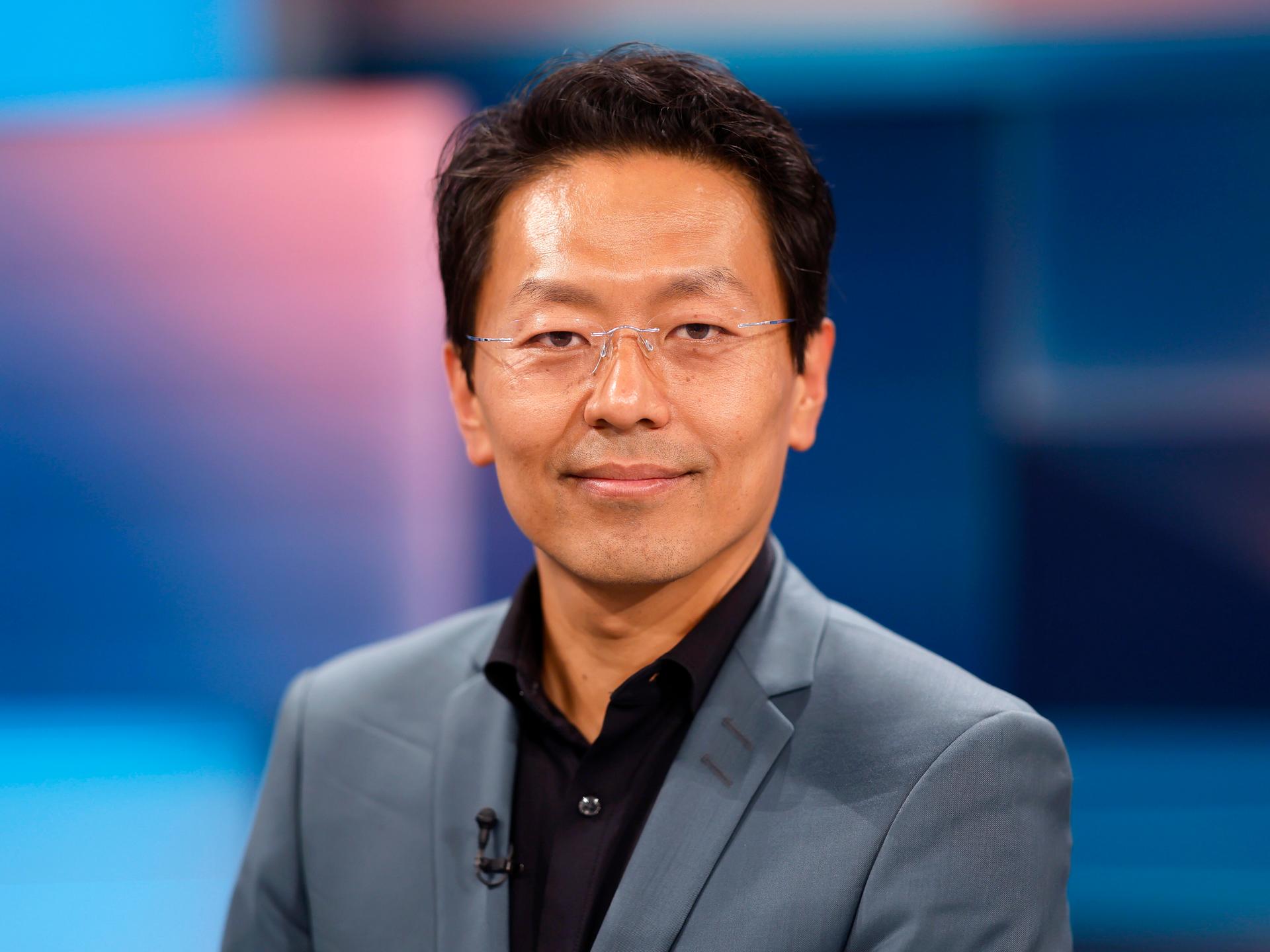 Jurist Chan-Jo Jun in den WDR-Studios in Köln. Er guckt in die Kamera. Er trägt eine rahmenlose Brille, ein schwarzes Hemd und einen grauen Anzug. Der Hintergrund ist bläulich.