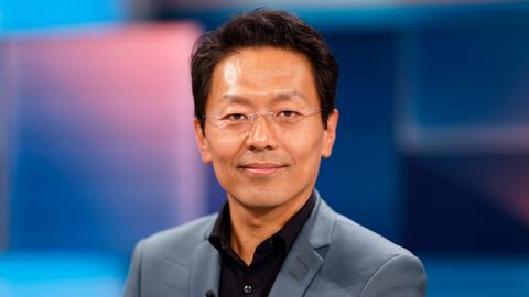 Jurist Chan-Jo Jun in den WDR-Studios in Köln. Er guckt in die Kamera. Er trägt eine rahmenlose Brille, ein schwarzes Hemd und einen grauen Anzug. Der Hintergrund ist bläulich.