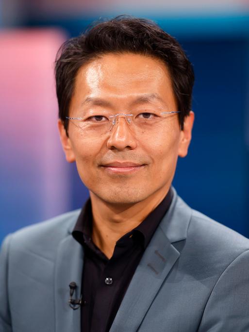 Jurist Chan-Jo Jun in den WDR-Studios in Köln. Er guckt in die Kamera. Er trägt eine rahmenlose Brille, ein schwarzes Hemd und einen grauen Anzug. Der Hintergrund ist bläulich.