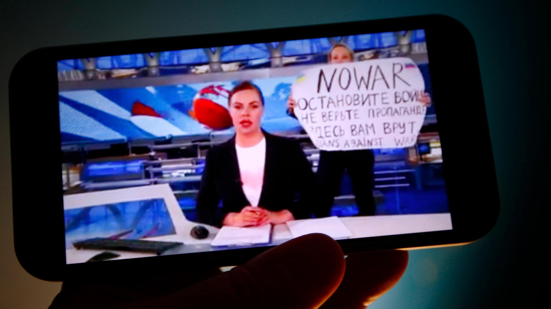 Auf dem Bildschirm eines Smartphones ist eine Nachrichtensprecherin zu sehen und hinter ihr eine Frau, die ein Schild mit der Aufschrift "No War" (Kein Krieg) hochhält.