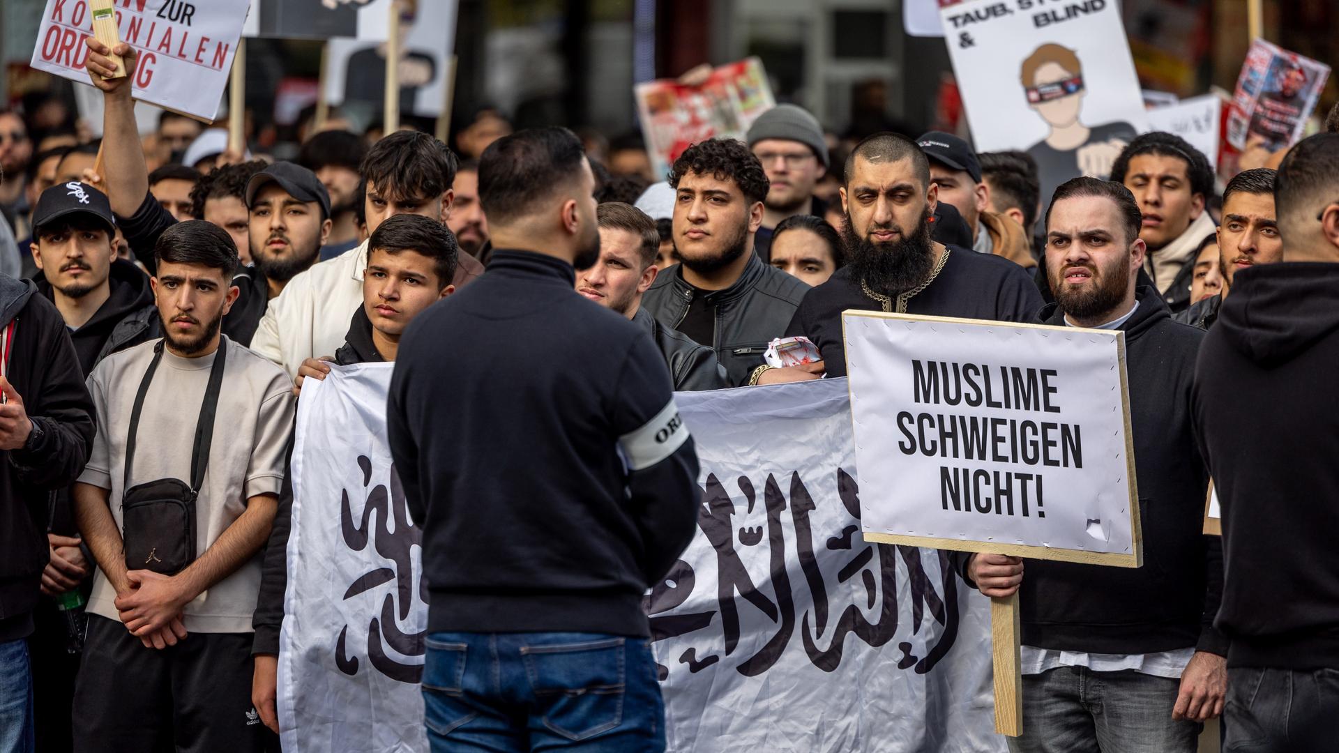 Ein Ordner steht mit dem Rücken zum Betrachter vor einer großen Menge an Demonstranten mit Transparenten, Spruchbändern und Schildern. Auf einem steht "Muslime schweigen nicht".