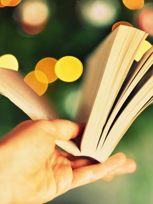 Eine Hand hält ein aufgeschlagenes Buch vor verschwommenem Hintergrund, der so aussieht, als sei es ein Weihnachtsbaum mit Beleuchtung.