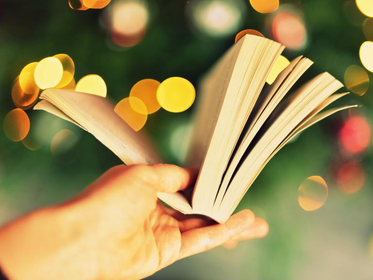 Eine Hand hält ein aufgeschlagenes Buch vor verschwommenem Hintergrund, der so aussieht, als sei es ein Weihnachtsbaum mit Beleuchtung.