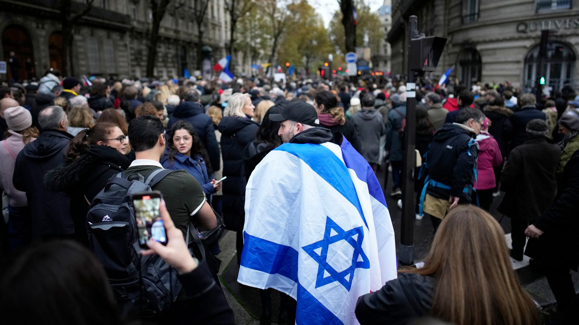 Das Bild zeigt eine große Menschenmenge, die durch eine Straße in der Innenstadt zieht. Im Bildzentrum ein Mann, der sich eine israelische Flagge umgehängt hat.