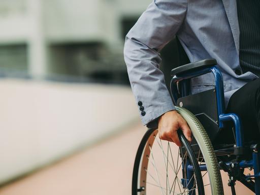 Ein Mann mit Sakko sitzt in einem Rollstuhl vor einem Geschäftsgebäude. Zu sehen ist nur seine Hand an einem Rad des Rollstuhls