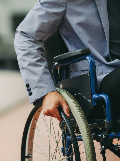 Ein Mann mit Sakko sitzt in einem Rollstuhl vor einem Geschäftsgebäude. Zu sehen ist nur seine Hand an einem Rad des Rollstuhls