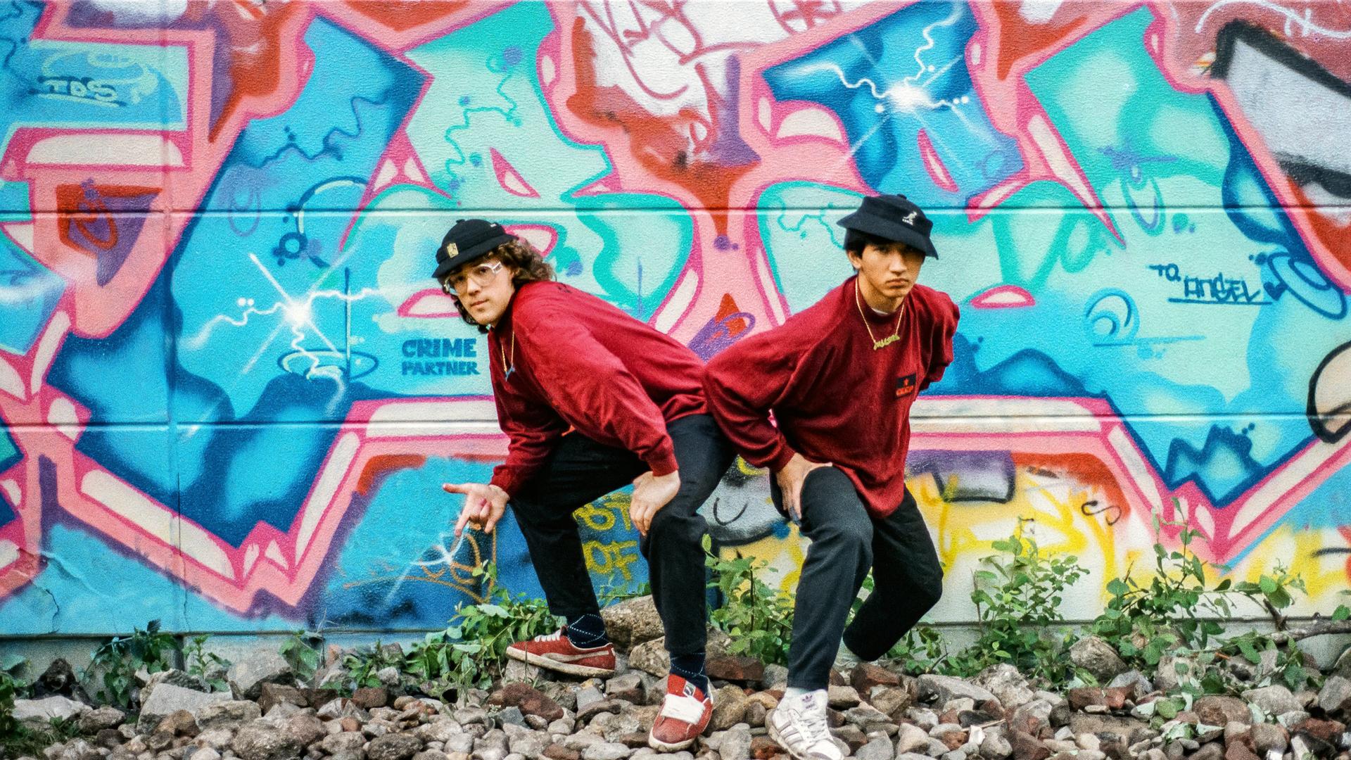 Zwei junge Männer posieren vor einem Graffiti.