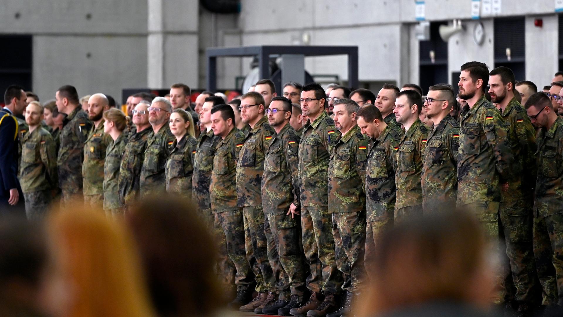 Soldaten stehen bei einem Verabschiedungsappell in Uniform in mehreren Reihen, im Vordergrund sieht man unscharfe Köpfe von hinten. 