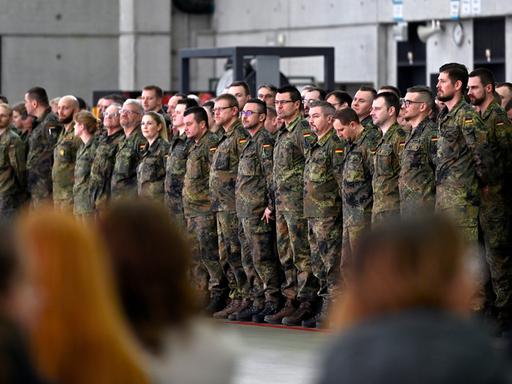 Soldaten stehen bei einem Verabschiedungsappell in Uniform in mehreren Reihen, im Vordergrund sieht man unscharfe Köpfe von hinten. 