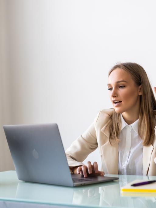 Eine junge Frau in Blazer und weißer Bluse sitzt an einem Büroschreibtisch vor einem Laptop. Sie scheint mit einer Person auf dem Bildschirm zu sprechen. Eine junge Frau in Blazer und weißer Bluse sitzt an einem Büroschreibtisch vor einem Laptop. Sie scheint mit einer Person auf dem Bildschirm zu sprechen.