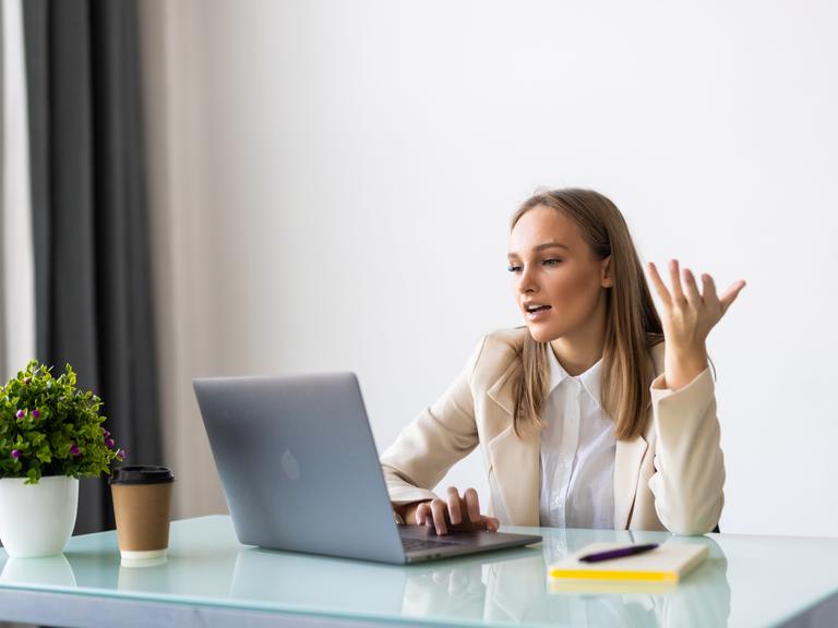 Eine junge Frau in Blazer und weißer Bluse sitzt an einem Büroschreibtisch vor einem Laptop. Sie scheint mit einer Person auf dem Bildschirm zu sprechen. Eine junge Frau in Blazer und weißer Bluse sitzt an einem Büroschreibtisch vor einem Laptop. Sie scheint mit einer Person auf dem Bildschirm zu sprechen.