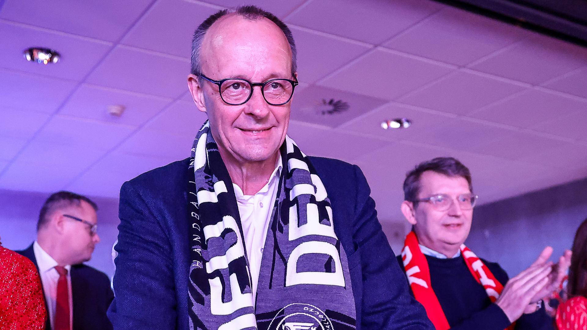 Bundeskanzler Friedrich Merz verfolgt ein Spiel mit Fanschal um den Hals.