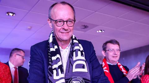 Bundeskanzler Friedrich Merz verfolgt ein Spiel mit Fanschal um den Hals.