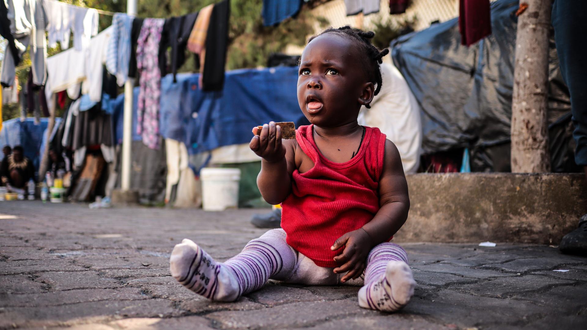 Ein kleines, dunkelhäutiges Mädchen sitzt in einem Flüchtlingslager auf dem Boden und hat einen dreckigen Gegenstand in der Hand. Im Hintergrund hängt Wäsche zum Trocknen. 