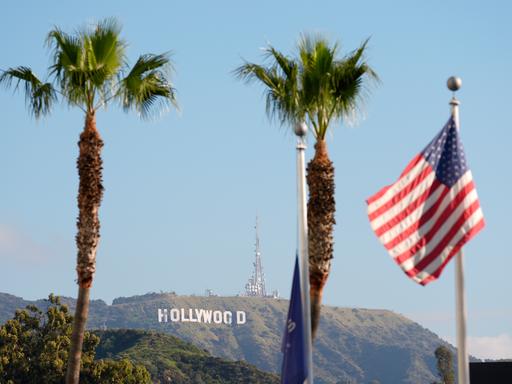 Im Hintergrund steht das Hollywood-Zeichen am Hang, im Vordergrund stehen Palmen und die US-Flagge hängt an einem Mast. Im Hintergrund steht das Hollywood-Zeichen am Hang, im Vordergrund stehen Palmen und die US-Flagge hängt an einem Mast.