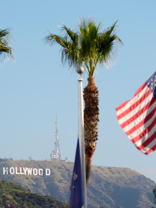 Im Hintergrund steht das Hollywood-Zeichen am Hang, im Vordergrund stehen Palmen und die US-Flagge hängt an einem Mast. Im Hintergrund steht das Hollywood-Zeichen am Hang, im Vordergrund stehen Palmen und die US-Flagge hängt an einem Mast.