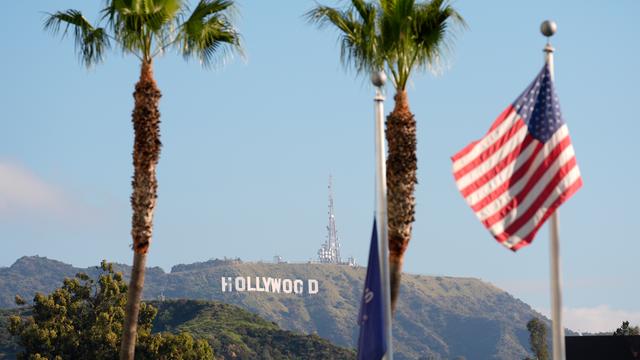 Im Hintergrund steht das Hollywood-Zeichen am Hang, im Vordergrund stehen Palmen und die US-Flagge hängt an einem Mast.