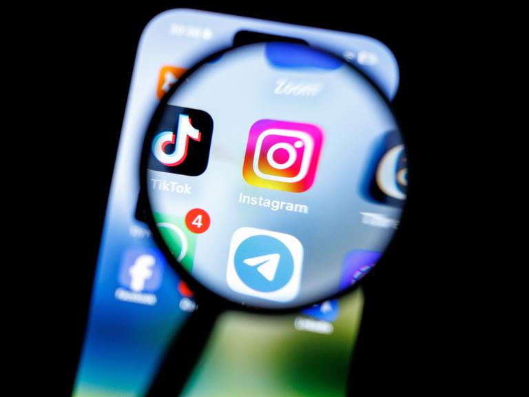 Das Logo der App Instagram ist durch eine Lupe auf dem Display eines Smartphones zu sehen. (Symbolbild)