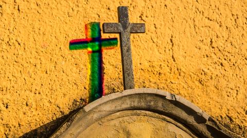 Kreuz auf einem Grabstein an einer gelben Wand mit Schatten in Regenbogenfarben 