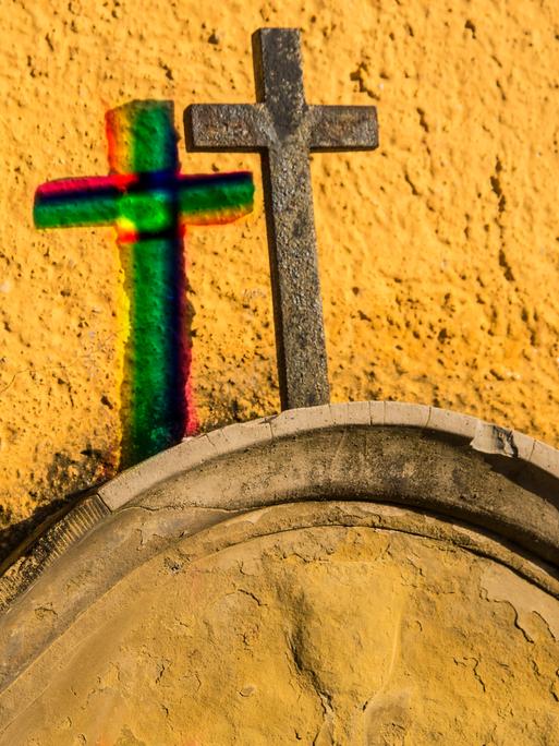Kreuz auf einem Grabstein an einer gelben Wand mit Schatten in Regenbogenfarben 