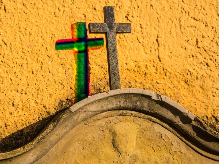 Kreuz auf einem Grabstein an einer gelben Wand mit Schatten in Regenbogenfarben 